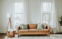 Salon avec uncanapé en cuir vintage, installé sur un tapis blanc, derrière lui 2 fenêtres avec des rideaux voilage, à gauche de la photo un luminaire sur pied vintage et un panier posé au sol avec un plaid dedans.