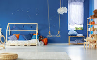 Chambre d'enfant peinte en bleu nuit, avec un lit cabane, une balançoire, un meuble à 5 étagère en bois, un tapis clair, un pouf en rotin et des accessoires dans le thème du ciel.