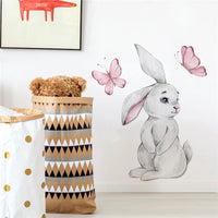 Sticker Mural Chambre avec un Lapin et Deux Papillons collé sur un mur blanc avec un tableau et deux sacs a jouet devant avec un ours en peluche dedans